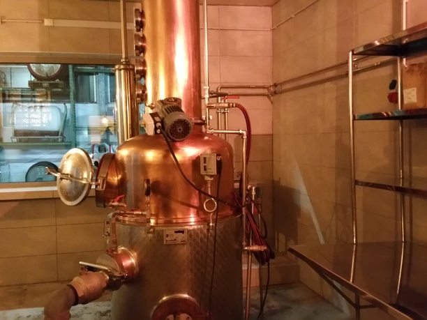 Kornbrennerei Hoffer Alter Kupferne Destillieranlage in einer Brennerei, umgeben von Fliesenwänden und einem Regal aus Metall.