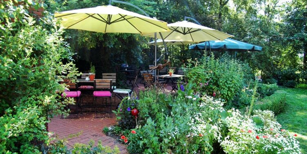 2013-07-21.jpg Ein idyllischer Garten mit üppigem Grün, Blumenbeeten und Sitzplätzen unter großen Sonnenschirmen.