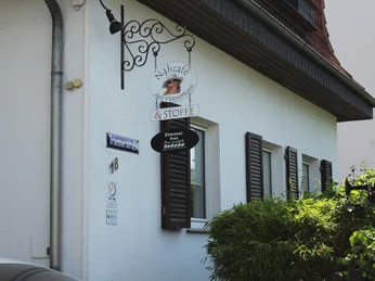 Nähcafé Nähcafé mit traditionellem Außenbereich, Holzläden, stilvollem Schild und grünen Pflanzen davor.