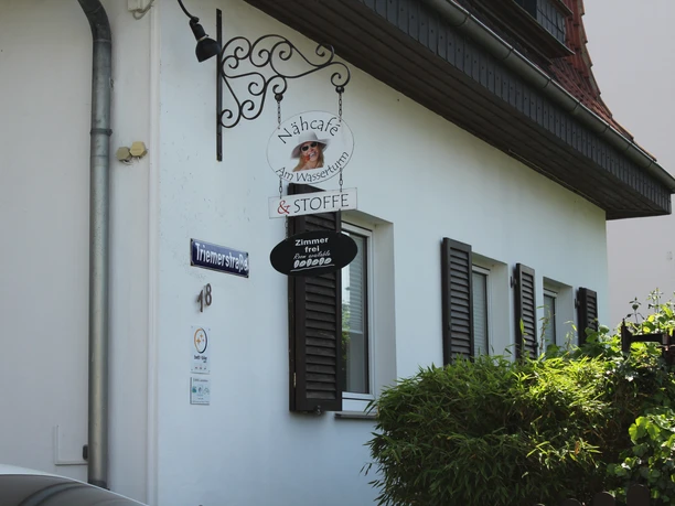 Nähcafé Nähcafé mit traditionellem Außenbereich, Holzläden, stilvollem Schild und grünen Pflanzen davor.