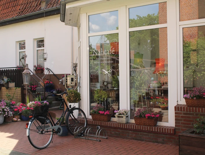 Nähcafé Einladendes Nähcafé mit blühenden Blumen und einem Fahrrad vor großen, einladenden Fenstern.