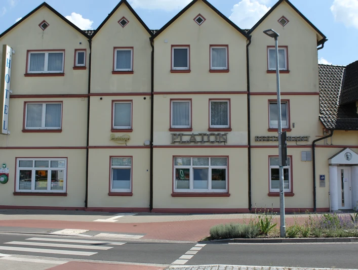 Drei-stöckiges Gebäude des Hotels Platon mit weißer Fassade und markant roten Fensterrahmen.Three-storey Hotel Platon building with a white façade and striking red window frames.Treetagers Hotel Platon-bygning med hvid facade og markante røde vinduesrammer.Hotel Platon van drie verdiepingen met een witte gevel en opvallende rode raamkozijnen.