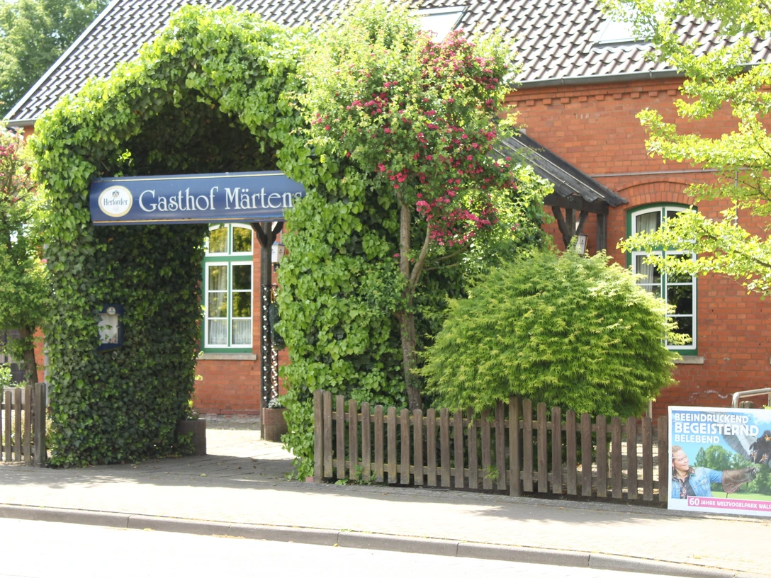 Historischer Gasthof mit üppiger Begrünung und einladendem Torbogen; rote Klinkerfassade im Hintergrund.