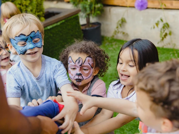 Kinder mit bunten Gesichtsbemalungen lachen und spielen fröhlich im Freien bei strahlendem Wetter.