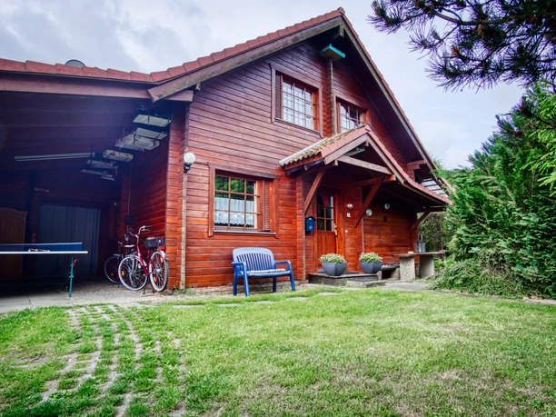 Rotbraunes Holzhaus mit grünem Vorgarten, Bank, Fahrrädern und einer Tischtennisplatte.