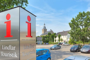 Tourist-Information Lindlar Ein Touristikinformationsschild in Lindlar, mit Kirche und parkenden Autos im Hintergrund.
