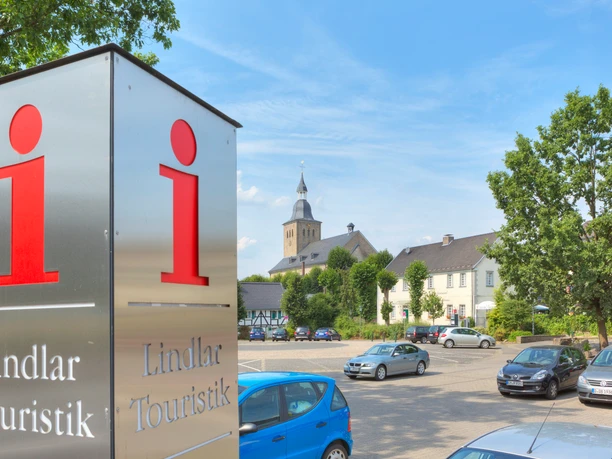 Tourist-Information Lindlar Ein Touristikinformationsschild in Lindlar, mit Kirche und parkenden Autos im Hintergrund.