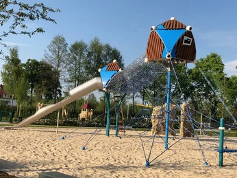 Ein moderner Spielplatz mit Klettergerüst und Rutsche auf Sandboden, umgeben von Bäumen im Freien.