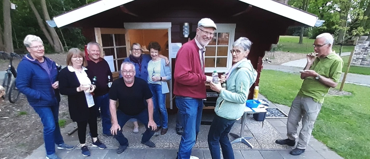 Eine Gruppe von acht Personen versammelt sich fröhlich bei einem Grillabend vor einer kleinen, rot gestrichenen Hütte. Im Vordergrund reicht ein Mann in einem roten Pullover einer Frau ein Getränk. Im Hintergrund befinden sich Bäume und Grünflächen, die eine gemütliche, naturnahe Atmosphäre schaffen.