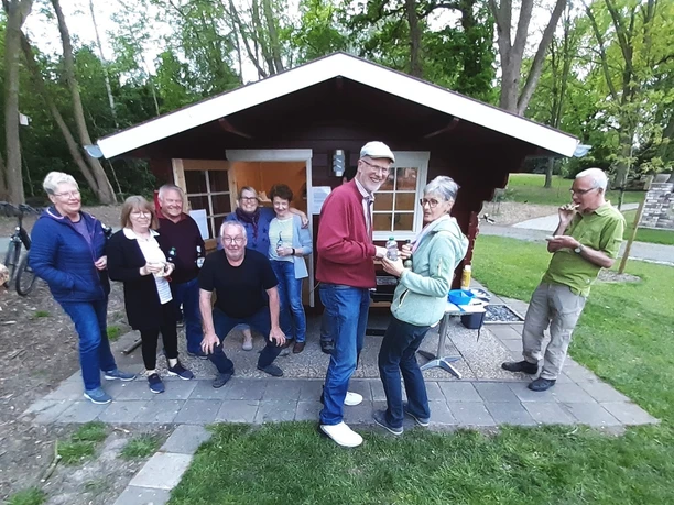 Grillabend Lüttje Festung Eine Gruppe von acht Personen versammelt sich fröhlich bei einem Grillabend vor einer kleinen, rot gestrichenen Hütte. Im Vordergrund reicht ein Mann in einem roten Pullover einer Frau ein Getränk. Im Hintergrund befinden sich Bäume und Grünflächen, die eine gemütliche, naturnahe Atmosphäre schaffen.