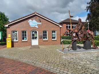 Apen Touristik mit Ritter Das Bild zeigt das Apen Touristik-Gebäude aus rotem Backstein mit einem blauen Schriftzug auf der Fassade. Daneben steht eine imposante Metallskulptur eines Ritters zu Pferd.