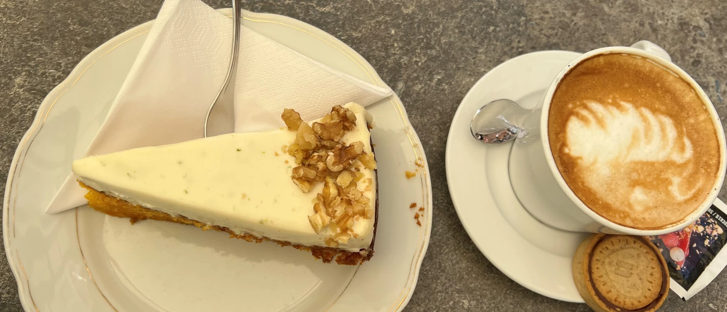 A slice of walnut cake served next to a cup of cappuccino with a cookie on a stone table.