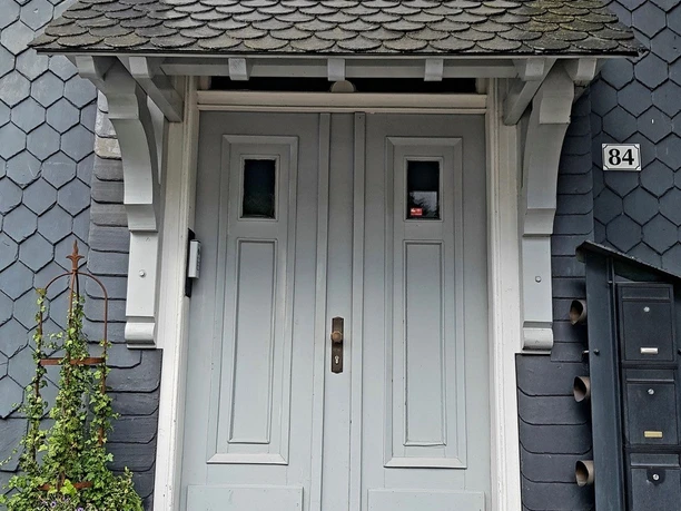 Eingang.jpg Doppeltür in Grau mit architektonischen Details und Laterne bewachsenen Blumenkübel daneben.