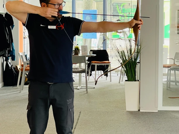 Ein Mann mit Brille zielt mit einem Bogen in einem modernen Innenraum. Helle Umgebung, Fokus auf Konzentration.
