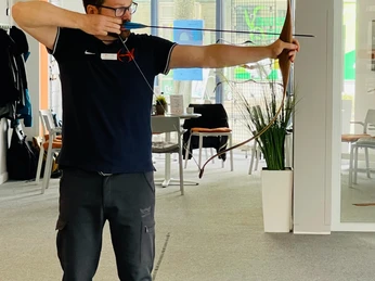 TBo-Laukert Ein Mann mit Brille zielt mit einem Bogen in einem modernen Innenraum. Helle Umgebung, Fokus auf Konzentration.