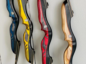 TBo-Laukert Bogen Vier hölzerne Recurvebögen in verschiedenen Farben hängen vertikal an einer Wand aus grauem Beton.