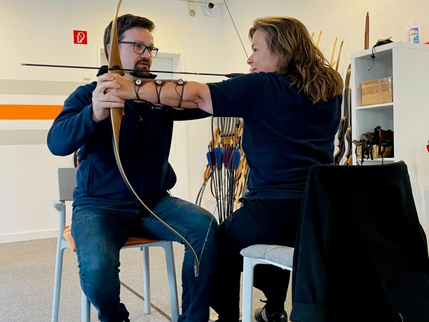 Zwei Personen sitzen drinnen, üben Bogenschießen mit traditionellem Bogen, Pfeile im Hintergrund.