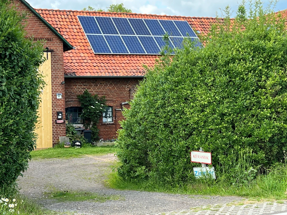 Das Bild zeigt ein rotes Backsteingebäude mit Solarpaneelen auf dem Dach, umgeben von grüner Vegetation.