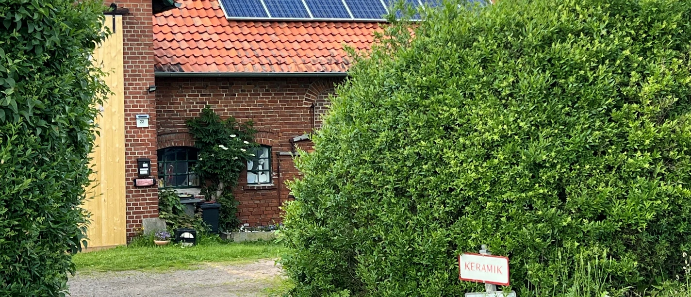 Das Bild zeigt ein rotes Backsteingebäude mit Solarpaneelen auf dem Dach, umgeben von grüner Vegetation.