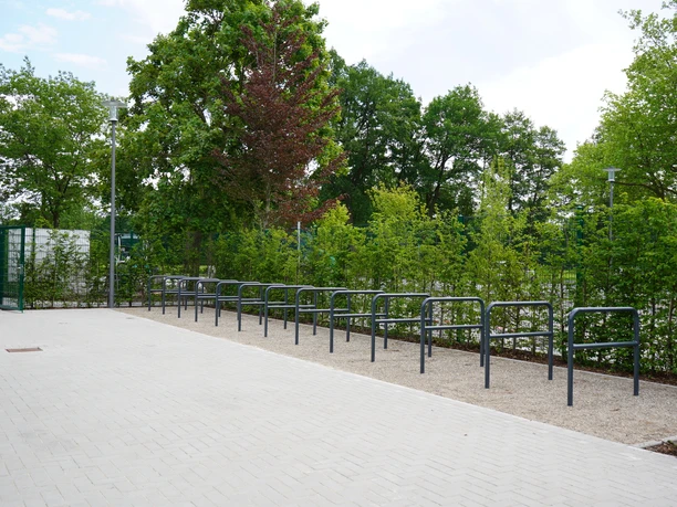 Reihe moderner Fahrradständer neben einem gepflasterten Weg, umgeben von grünen Bäumen und Büschen.