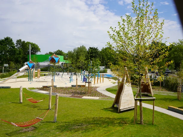 Moderner Spielplatz mit Klettergerüsten, Rutschen und Hängematten, umgeben von grüner Landschaft.