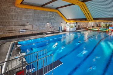 Parkbad Innenansicht eines Hallenbads mit blauem Schwimmbecken, Sprungturm und Badegästen im Wasser.