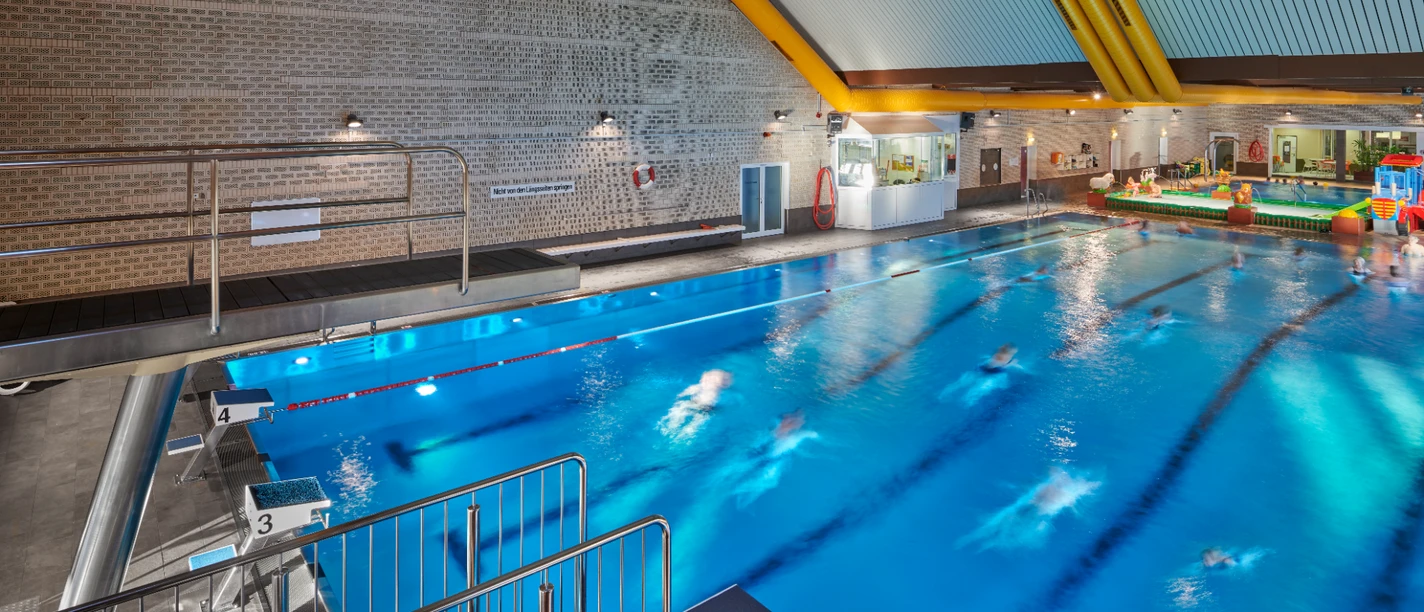 Parkbad Innenansicht eines Hallenbads mit blauem Schwimmbecken, Sprungturm und Badegästen im Wasser.