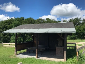 Grillhütte Kapellensüng_Schlüsselberg Überdachte Holzhütte mit Picknicktisch und Grillplatz auf einer grünen Wiese unter blauem Himmel.