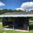 Grillhütte Kapellensüng_Schlüsselberg Überdachte Holzhütte mit Picknicktisch und Grillplatz auf einer grünen Wiese unter blauem Himmel.