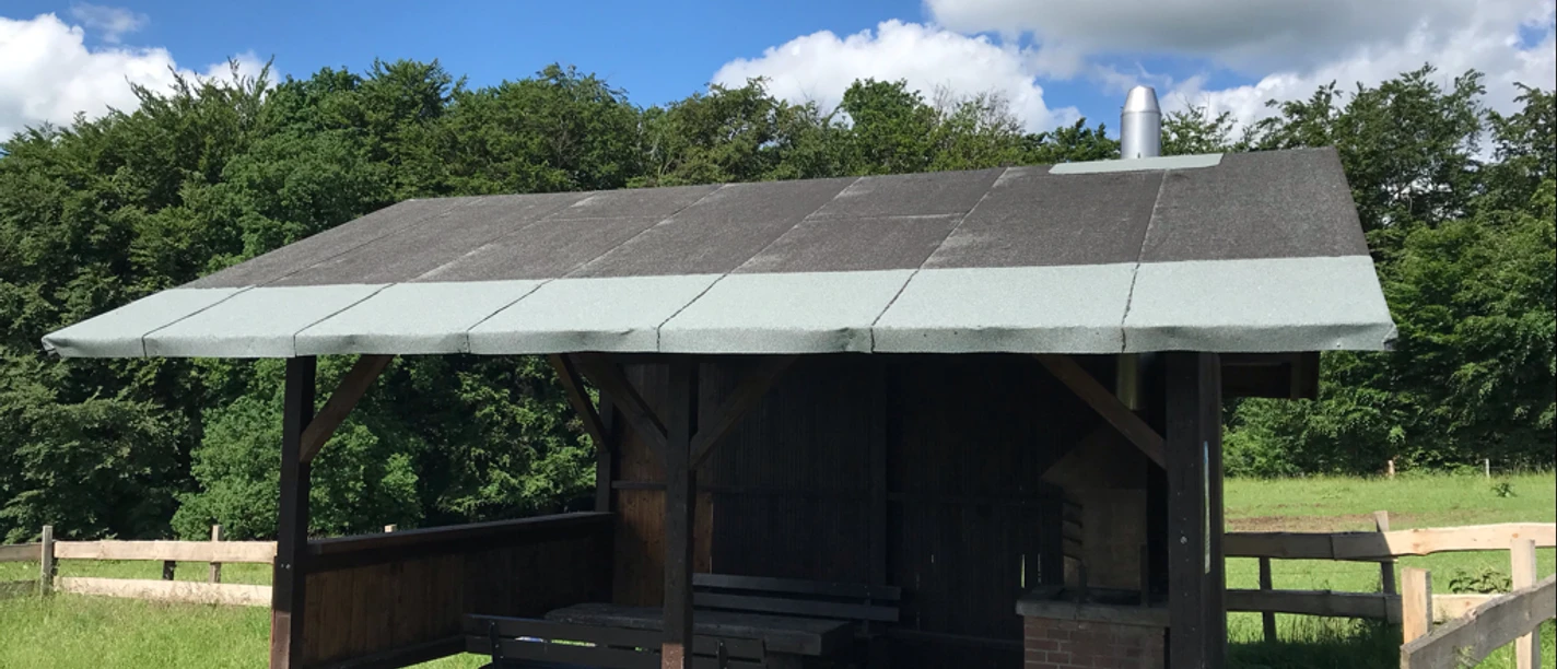 Grillhütte Kapellensüng_Schlüsselberg Überdachte Holzhütte mit Picknicktisch und Grillplatz auf einer grünen Wiese unter blauem Himmel.