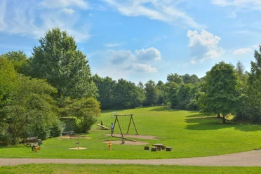 Freizeitpark (Grillplatz) Ein Spielplatz mit Schaukeln und Grünflächen in einem sonnigen Park mit blauen Himmel und Bäumen.