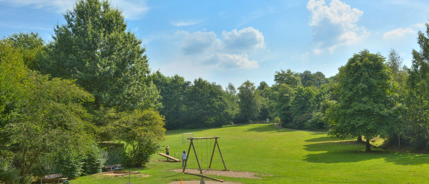 Freizeitpark (Grillplatz) Ein Spielplatz mit Schaukeln und Grünflächen in einem sonnigen Park mit blauen Himmel und Bäumen.
