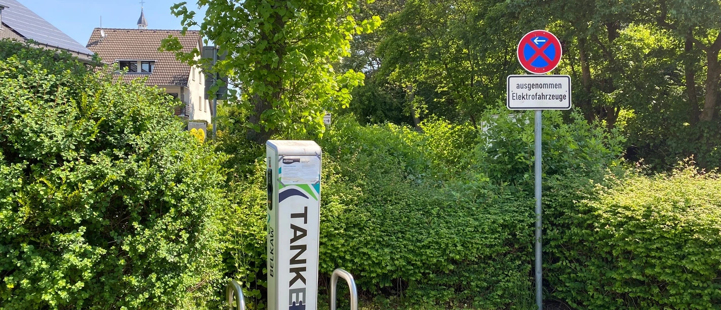 Ladesäule Kölner Straße Ladesäule für Elektrofahrzeuge zwischen grünen Hecken, daneben Parkverbots- und Reserviert-Schild.