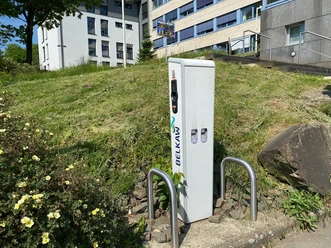 Ladesäule Rathaus Ladestation von BELKAW für E-Bikes vor einem modernen Gebäude mit grüner Rasenfläche und Pflanzen.