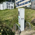Ladesäule Rathaus Ladestation von BELKAW für E-Bikes vor einem modernen Gebäude mit grüner Rasenfläche und Pflanzen.