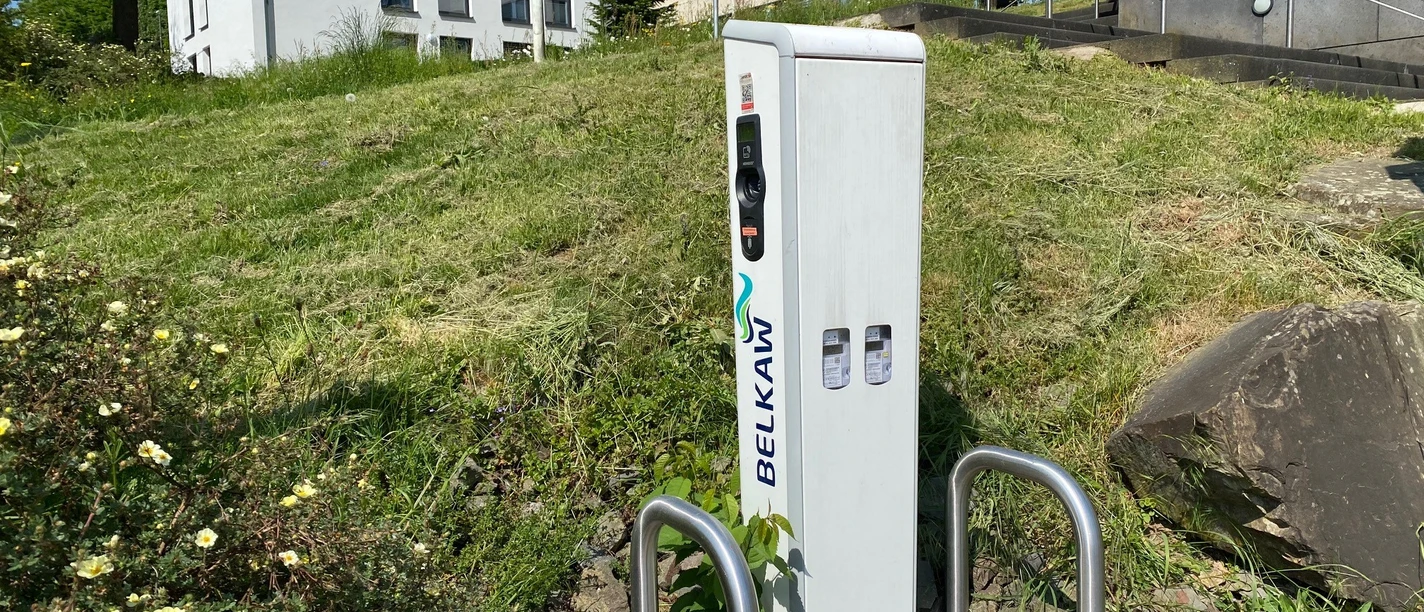 Ladesäule Rathaus Ladestation von BELKAW für E-Bikes vor einem modernen Gebäude mit grüner Rasenfläche und Pflanzen.