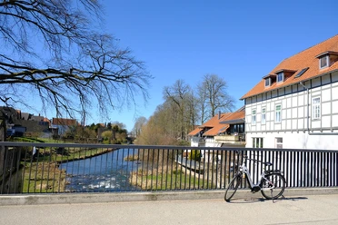 E-Bike an der Bega.JPG Ein E-Bike lehnt an einer Brücke über einen Fluss, umgeben von Fachwerkhäusern. Klarer Himmel.
