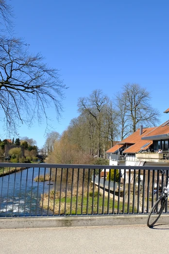 E-Bike an der Bega.JPG Ein E-Bike lehnt an einer Brücke über einen Fluss, umgeben von Fachwerkhäusern. Klarer Himmel.