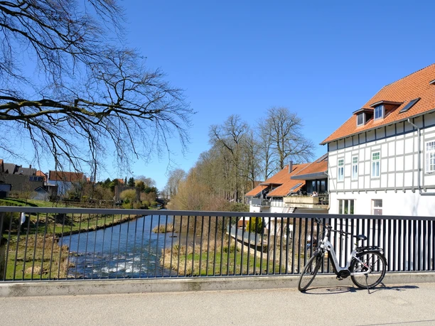 E-Bike an der Bega.JPG Ein E-Bike lehnt an einer Brücke über einen Fluss, umgeben von Fachwerkhäusern. Klarer Himmel.