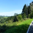 Bahntrasse Sülztalbahn©Lindlar Touristik.JPG Weg führt durch bewaldetes Gebiet mit weitem Blick auf bewachsene Hügel unter strahlend blauem Himmel.