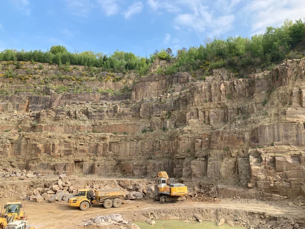 Blick in einen Steinbruch Steinbruch mit Bagger und Lastwagen vor beeindruckender Felswand unter blauem Himmel mit Wolken.