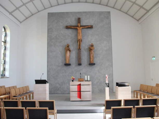 Schlichter Kircheninnenraum mit Altar, Kruzifix und Figuren an einer grauen Wand, helle Holzbänke.