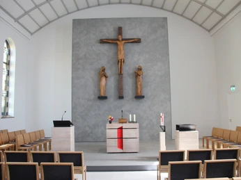 Kreuzkirche Wiedenbrück Schlichter Kircheninnenraum mit Altar, Kruzifix und Figuren an einer grauen Wand, helle Holzbänke.