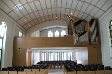 Moderner Kirchenraum mit geschwungenem Dach, Orgel links, hellem Holz, leeren Stuhlreihen und Fenstern.