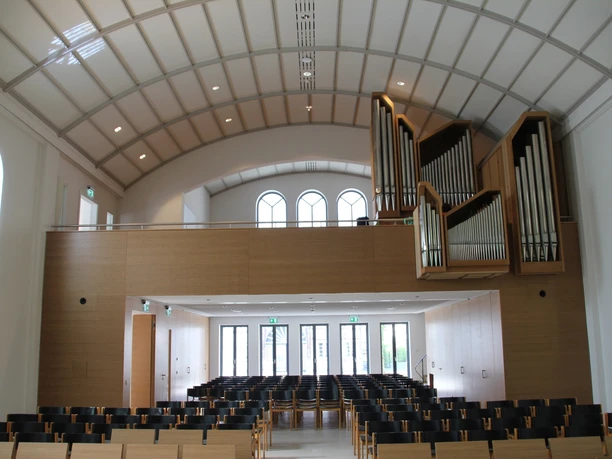 Moderner Kirchenraum mit geschwungenem Dach, Orgel links, hellem Holz, leeren Stuhlreihen und Fenstern.