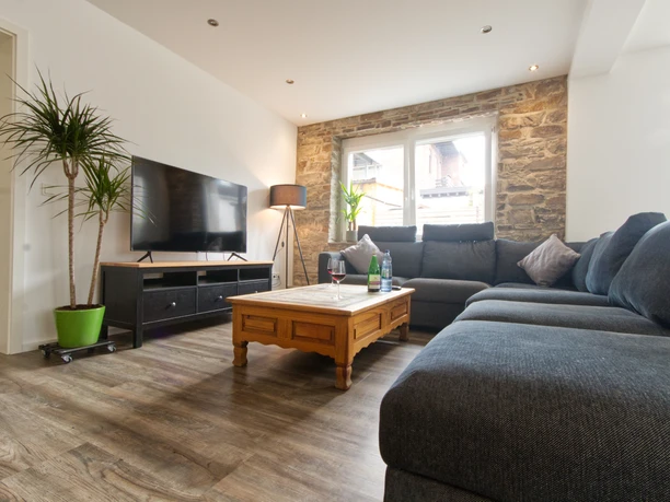 Ferienwohnung Wiehl Wohnzimmer.jpg Gemütliches Wohnzimmer mit grauem Ecksofa, Holztisch, Pflanzen und Wandfernseher vor Steinwand.