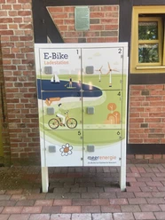 Ladestation für E-Bikes mit sechs Schließfächern vor roter Backsteinwand mit Holzrahmenfenster.
