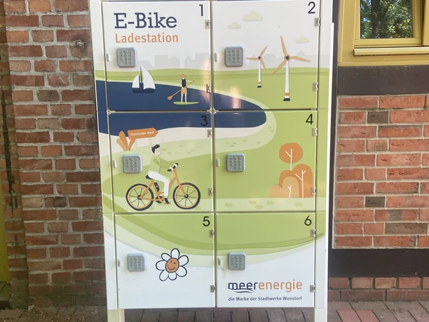 Ladestation für e-bikes Ladestation für E-Bikes mit sechs Schließfächern vor roter Backsteinwand mit Holzrahmenfenster.