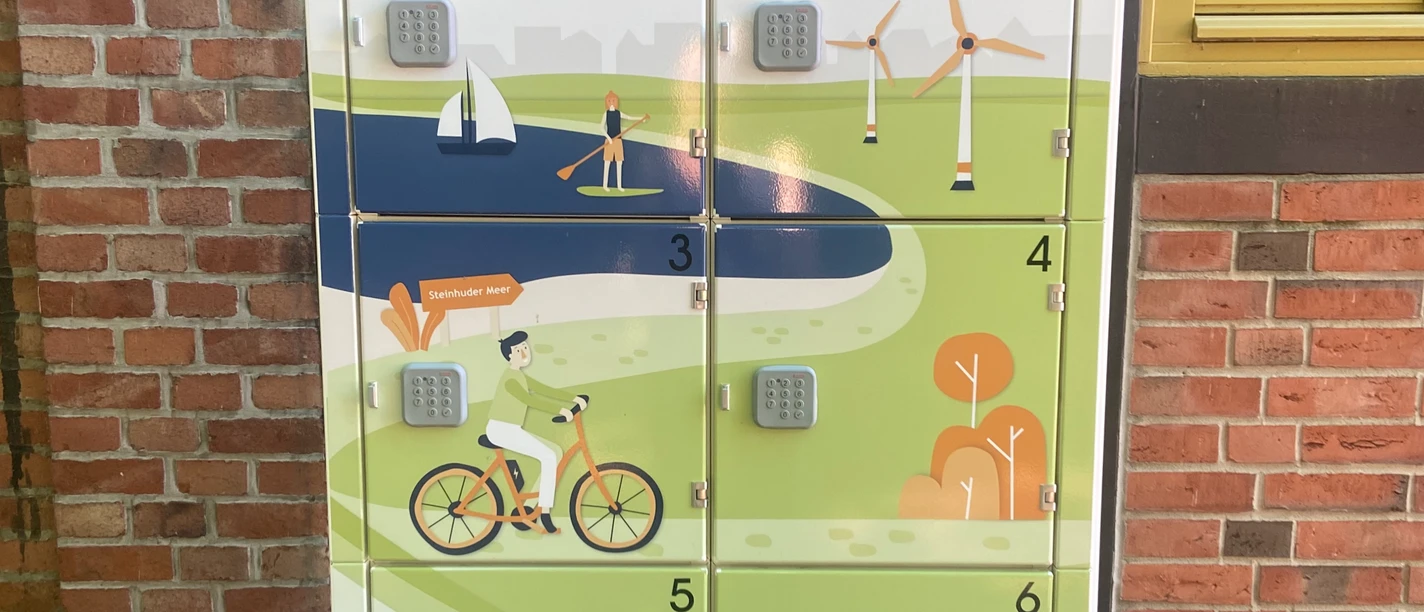 Ladestation für E-Bikes mit sechs Schließfächern vor roter Backsteinwand mit Holzrahmenfenster.