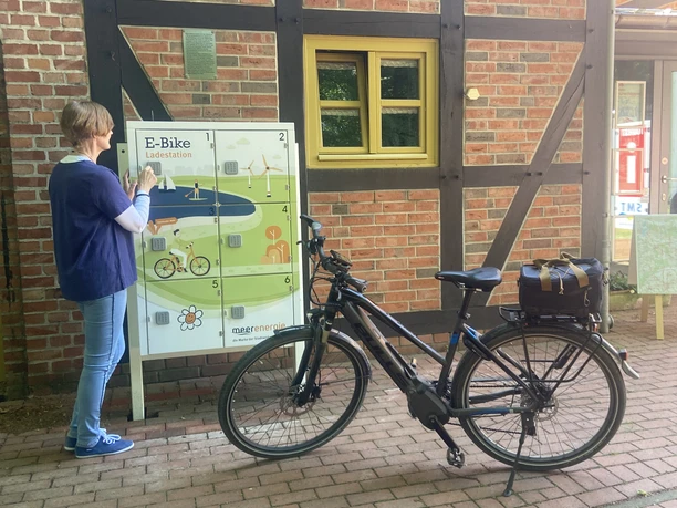Eine Person bedient eine E-Bike-Ladestation an einer Fachwerkfassade, ein E-Bike steht daneben.
